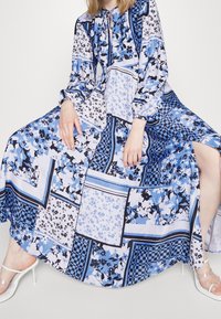 Lange blauwe jurk met bloemenprint en patroon van vierkanten, voorzien van een geraffineerde halslijn, lange mouwen en een zijsplit. Draagt heldere sandalen met hak.