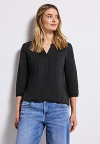 Femme aux cheveux blonds mi-longs portant un blouse noire à boutons avec des manches 3/4 et un jean en denim bleu, debout contre un mur blanc.