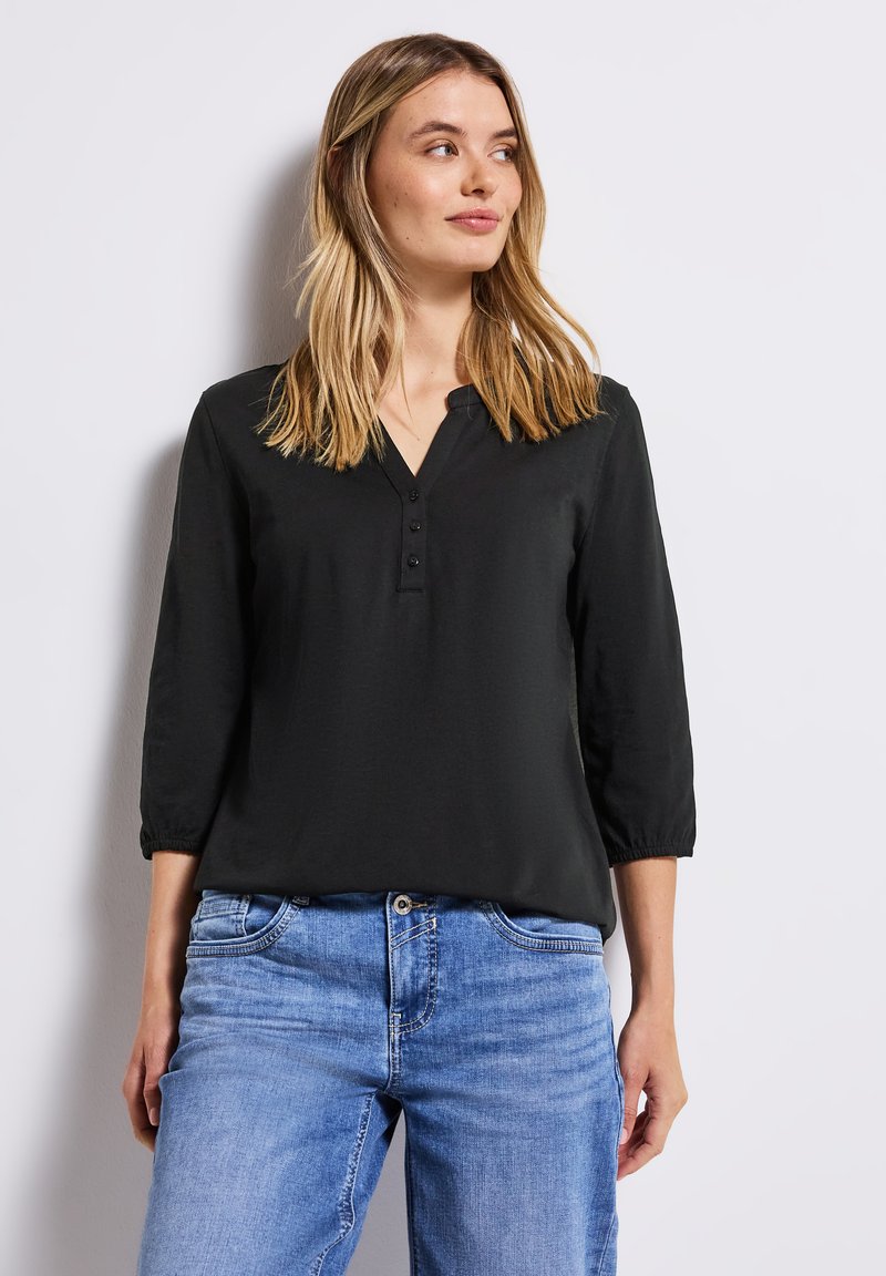 Femme aux cheveux blonds mi-longs portant un blouse noire à boutons avec des manches 3/4 et un jean en denim bleu, debout contre un mur blanc.