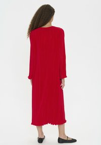 Rochie roșie plisată, cu mâneci lungi, decolteu rotund și tiv volanat. Purtați-o cu pantofi negri de tip plat, cu un design minimalist.