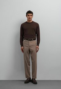 Chemise marron à manches longues, pantalon beige, ceinture noire et chaussures marron foncé, debout devant un fond neutre. Design minimaliste.