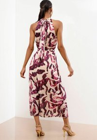 Donna che indossa un vestito midi senza maniche, annodato sul retro, con un motivo in tonalità di rosa e viola, abbinato a sandali con tacco dorati, in piedi su uno sfondo bianco.