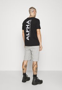 Crna kratka majica s kratkim rukavima s bijelim logotipom "ALPHA INDUSTRIES" na leđima, uparena s sivoj kratkim hlačama i crnim čizmama na vezivanje.