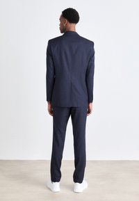 Completo uomo navy con design su misura, dotato di colletto alto e pantaloni slim-fit. Indossato con sneakers bianche. Texture liscia.