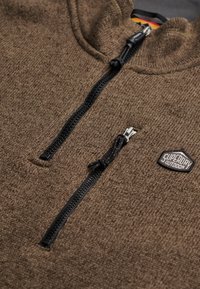 Bruine fleece met ritssluiting en een gestructureerde afwerking, voorzien van een zwarte rits en een patch met daarop "Superdry Outdoor" op de borst.