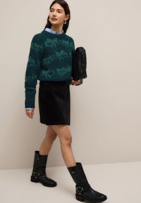 Zeleno-modri debeli pleteni pulover z vzorcem pentlje, črna kordurova krila ter črni usnjeni škornji do kolen z naramnicami in srebrnimi poudarki.