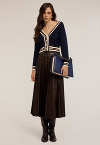 Námořnický pletený kardigan s krémovými pruhy, s knoflíkovými uzávěry, spárovaný s plisovanou hnědou koženou sukní a černými kozačkami po kolena, držící modrou tašku.