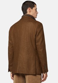 Cappotto di lana marrone con una finitura testurizzata, design su misura e colletto visibile. Presenta due bottoni sulla manica. Linee pulite, dettagli minimi.