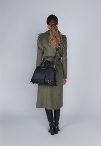 Cappotto lungo verde oliva con cintura, abbinato a una borsa nera con frange. La modella indossa stivali neri alti e ha i capelli ondulati raccolti.