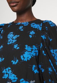 Detailní záběr na osobu, která má na sobě černou blůzu s jasně modrými květinovými vzory a sklady na balónových rukávech.