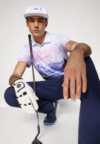 Mladý muž v bielej čiapke a golfovej rukavici, ktorý sa squattuje a drží golfovú palicu. Má na sebe pastelový polo s horským motívom a námornícke nohavice.