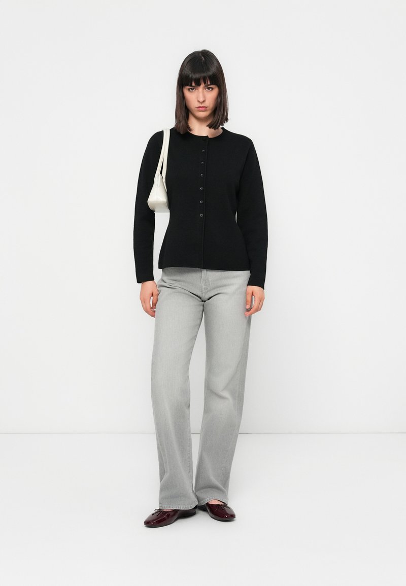 Czarny sweter zapinany na guziki, szare dżinsy typu wide-leg i bordowe buty. Nosi małą białą torbę. Prosty design i dopasowany krój.