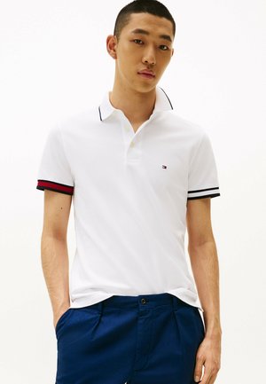 Maglietta polo bianca con colletto, caratterizzata da accenti a righe blu navy e rosse sulle maniche. Indossata con pantaloni blu navy. Design e texture semplici.
