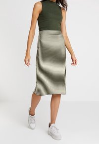 Pletený top bez rukávů v tmavě zelené barvě, kombinovaný s proužkovanou midi sukní v krémové a černé barvě. Nošeno s bílými teniskami.