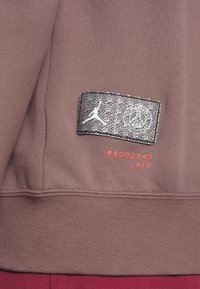 Brun sweatshirtstof med en tekstureret overflade. Har et sort og sølv patch med et logo, nummereret "#002345," og orange tekst.