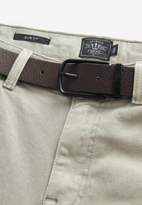 Pantalones ajustados beige con un cinturón de cuero marrón oscuro texturizado y hebilla de metal negro, que presenta una etiqueta con el logo de "Premium" y "next".