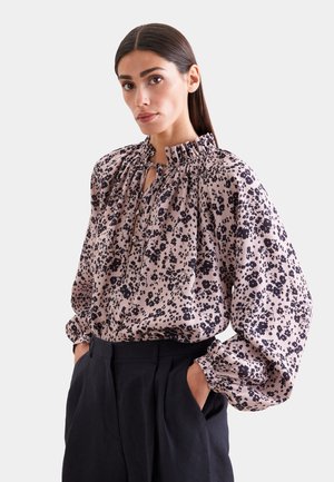 Kobieta z prostymi ciemnymi włosami, ubrana w różową kwiecistą bluzkę z długim rękawem i falbanianym kołnierzem oraz czarne spodnie z wysokim stanem, ręce w kieszeniach.