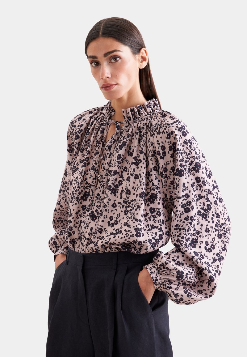 Femme aux cheveux raides et foncés portant un chemisier rose à fleurs à manches longues avec un col à volants et un pantalon noir taille haute, mains dans les poches.