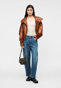 Brun läderjacka med bred krage, ljus linne, blå lösa jeans, grön väska och svarta skor. Modellen står mot en neutral bakgrund.