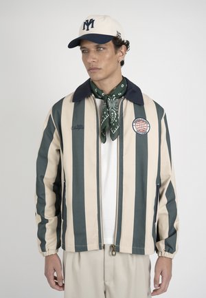 Mand iført creme og grøn lodretstribet jakke, hvid skjorte, grøn mønstret tørklæde, cremefarvet kasket med navy logo og beige bukser mod en ensfarvet baggrund.