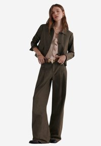 Olivengrüne taillierte Jacke mit Kragen, kombiniert mit einer hellrosa Rüschbluse und einer weiten Hose, die mit einem kontrastierenden Gürtel versehen ist.