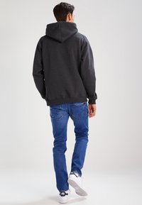 Kullsvart hettegensere med snøring, kombinert med blå jeans og hvite joggesko. Hettegenseren har en avslappet passform og ribbede mansjetter.