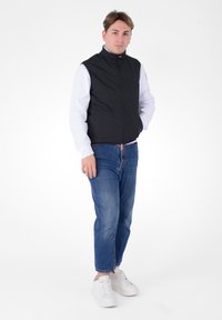 Gilet imbottito nero con colletto alto, chiusura con zip e tasche laterali; indossato sopra una maglietta bianca a maniche lunghe e abbinato a jeans blu e sneaker bianche.