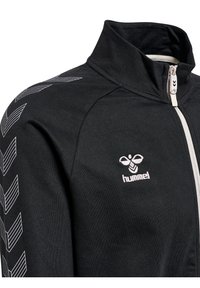 Giacca nera con zip realizzata in tessuto liscio. Presenta motivi a chevron bianchi sulle maniche e un logo sul petto. Design con colletto alto.