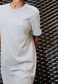 Person som bär en grå Adidas t-shirt med vita ränder på axlarna, stående mot en texturerad svart metallbakgrund.