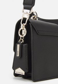 Musta tekokuituinen käsilaukku, jossa on kultasävyinen metalliketju ja brändätty tunnus. Laukussa on läppä suljenta ja muotonsa pitävä rakenne.