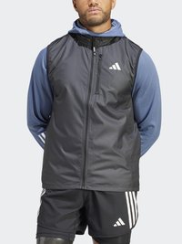 Svart idrettsvest med glidelås og hette, med en jevn tekstur og hvitt logo. Brukt over en blå langermet topp, med svarte shorts under.