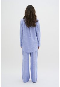 Cămașă albastră deschis, cu carouri, cu mâneci lungi și pantaloni largi asortați, amândouă dintr-un material moale, texturat. Manșetele cu nasturi sunt vizibile.