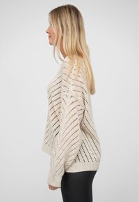 Offenes, gestricktes cremefarbenes Sweatshirt mit Zickzackmuster, weiten Ärmeln und lässiger Passform. Die strukturierte Materialbeschaffenheit steht im Kontrast zu den glatten schwarzen Leggings.
