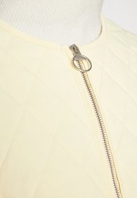 Veste jaune matelassée avec une texture lisse, dotée d'une fermeture éclair argentée avec un tirette rond, motifs en losange visibles, et un col côtelé.