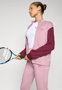 Ung kvinne i rosa og burgunder treningsdress som holder en tennisracket og ser til siden mot en ensfarget lys bakgrunn.