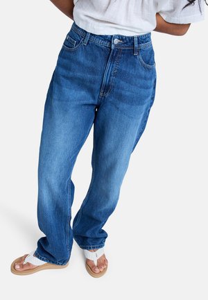 Frau trägt locker sitzende, hoch taillierte blaue Jeans mit weißen gewebten Sandalen, steht vor einem einfarbigen Hintergrund.