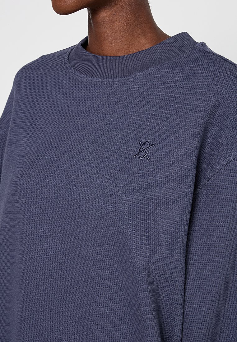 Donkerblauwe trui met wafeltextuur en een geribbelde ronde hals. Kenmerkt zich door een klein geborduurd logo op de borst. Zacht materiaal en een losse pasvorm.