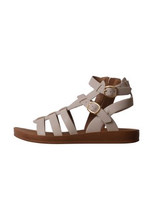 Sandale gladiateur plate beige avec semelle marron, plusieurs brides et deux fermetures à boucles dorées à la cheville et au pied.
