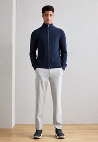 Navy zip-up sweater, slim-fit light gray trousers, and black sneakers. Smooth knit texture, minimalist design with ribbed cuffs and hem.