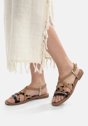 Füße in flachen Sandalen mit offenem Zehenbereich, Riemen in Braun, Schwarz und Leopardenmuster, kombiniert mit einem cremefarbenen, fransigen Rock bis zum Knöchel.