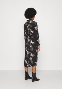 Abito nero floreale con maniche lunghe, realizzato in tessuto morbido. Presenta un fiocco al collo, motivi colorati e un taglio a lunghezza intermedia. Indossato con stivali neri alla caviglia.