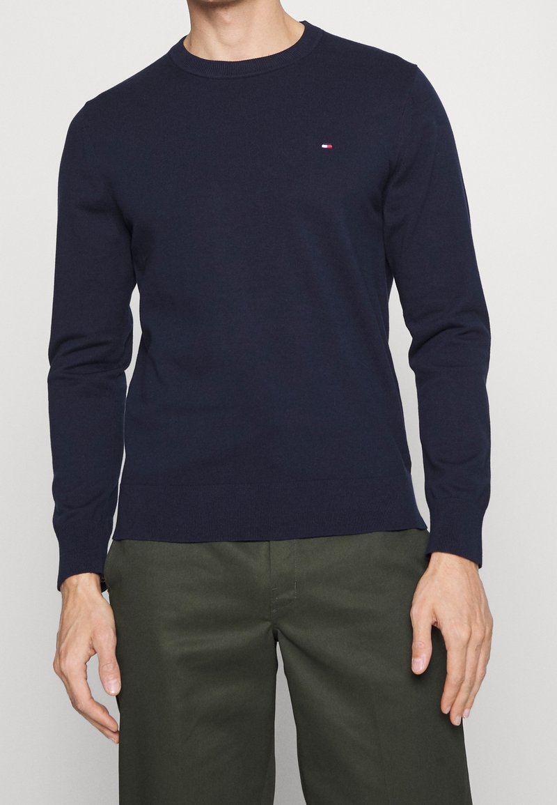 Homme portant un pull bleu marine à manches longues avec un petit logo rouge, blanc et bleu sur la poitrine et un pantalon vert olive.