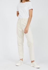 Pantalones de crema claro de talle alto con piernas ajustadas, hechos de una mezcla de algodón. Combinados con una camiseta blanca de manga corta y zapatillas blancas.
