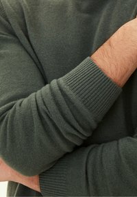 Maglione verde scuro con polsini a coste e tessuto texture, mostrato con un braccio piegato su uno sfondo neutro.