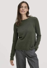 Frau in einem olivgrünen langärmeligen Pullover und grauen gestreiften Hosen, die mit einer Hand in der Tasche steht.