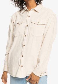 Beige kordfløjlsskjorte med krave, to brystlommer og brune knapper. Lange ærmer med knapmanchetter og ribbet tekstur.