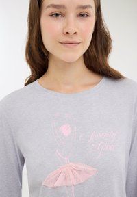 Giovane donna che indossa una maglietta grigia chiara con una schizzo di ballerina rosa caratterizzato da un tutù testurizzato e il testo "Amazing Grace".