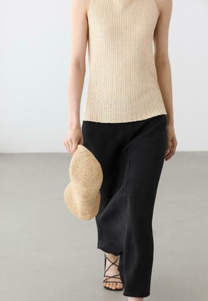 Personne portant un haut en tricot beige sans manches et un pantalon large noir, tenant un chapeau tissé, marchant sur un sol gris clair.