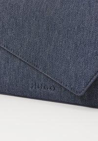Denim konvoluttlommebok i mørk blå, med teksturert overflate, synlig søm og preget logo på fronten.