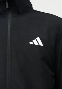 Melna kapuce jaka ar priekšējo rāvējslēdzēju un baltu trīsstūrveida logotipu kreisajā krūšu rajonā.